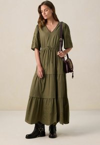 Robe en coton vert olive, longueur genou, avec un décolleté en V, des manches bouffantes et une taille à cordon. Elle présente une jupe à volants et des broderies.