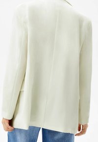 Osoba v dlouhém rukávu, off-white blazeru a modrých džínách, viděná zezadu s uvolněnýma rukama podél těla.