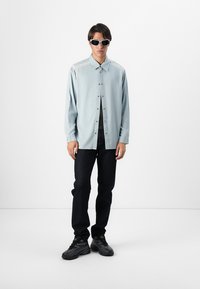 Svetlomodrá košeľa na gombíky, čierne džínsy a čierne tenisky. Model nosí elegantné biele slnečné okuliare a stojí na jednofarebnom pozadí.