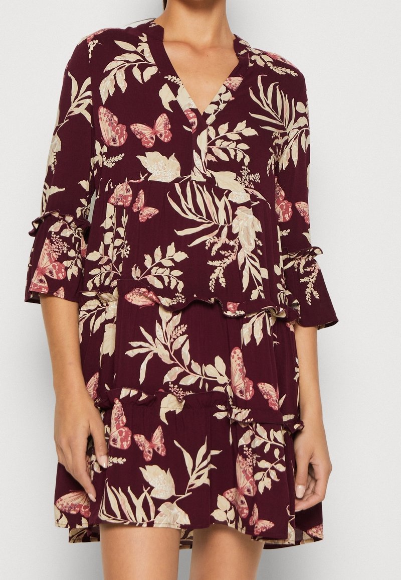 Femme portant une robe bordeaux avec des motifs floraux crème et des papillons roses, manches trois-quarts, et une jupe à étages avec volants.