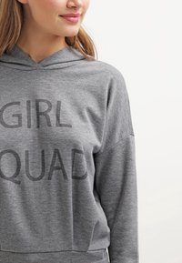 Sudadera con capucha gris con el texto "GIRL SQUAD" impreso en gris oscuro. Presenta un ajuste holgado y una tela suave y texturizada.