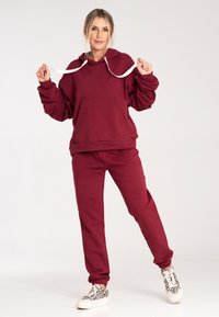 Bordowa bluza z kapturem z białymi sznurkami i przednią kieszenią, połączona z pasującymi bordowymi dresami. Posiada luźny krój i ściągacze przy rękawach.