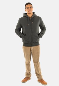 Haut à capuche zippé vert foncé avec des cordons, poches avant et poignets côtelés, associé à un pantalon en velours côtelé beige et des chaussures marron.