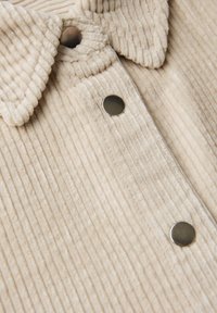 Beige corduroy overhemd met een insnoerboord, verticale ribbeltextuur en metalen drukknoopjes aan de voorkant.
