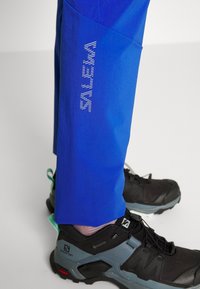 Siniset aktiivihousut, joissa on heijastava valkoinen logo reidessä, valmistettu kevyestä kankaasta, yhdistettynä mustiin vaelluskengiin.