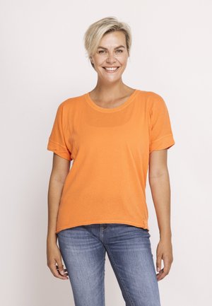 Lächelnde Frau mit kurzen blonden Haaren, die ein oranges Kurzarmshirt und blaue Jeans trägt, vor einem schlichten hellen Hintergrund stehend.