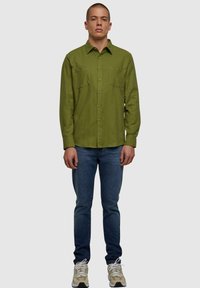 Camisa verde de manga larga con cierre de botón, presenta dos bolsillos en el pecho. Combinada con jeans azules y zapatillas beige sobre un fondo neutro.