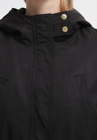Black hooded jacket featuring a zip closure, two side pockets, and gold snap buttons on the collar. Made of smooth, matte fabric.