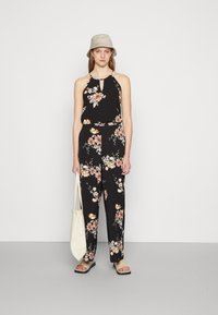 Jardineira preta com estampa floral e decote halterneck, apresentando flores em tons de rosa e creme. A modelo segura uma mala tote branca, usando sandálias pretas e um chapéu de palha bege.