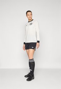 White long-sleeve soccer jersey with black collar and logo, paired with black shorts, black cleats, and knee-high black socks with white stripes.