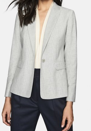Frau trägt einen hellgrauen Blazer mit einem Knopf über einer weißen Bluse und dunkelblauen Hosen, stehend vor einem schlichten Hintergrund.