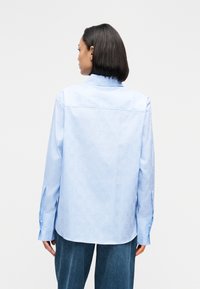 Világoskék, gombos ing finom mintával, hosszú ujjakkal és galléros nyakkivágással. Testre szabott szabású, mandzsetta gombokkal és lekerekített aljjal.