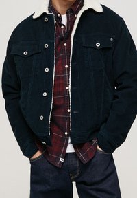 Donkerblauwe ribfluwelen jas met schapenvachtkraag, voorzien van twee borstzakken en zilverkleurige drukknopen, gedragen over een rood geruit overhemd.
