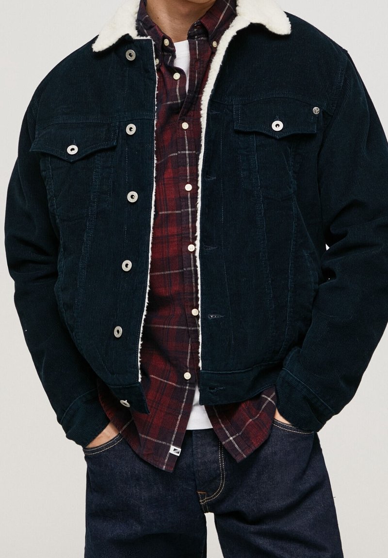 Donkerblauwe ribfluwelen jas met schapenvachtkraag, voorzien van twee borstzakken en zilverkleurige drukknopen, gedragen over een rood geruit overhemd.