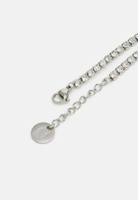 Silberne Kettenarmband mit klaren Strasssteinen, Karabinerverschluss und einem runden Logo-Anhänger. Glatte Textur mit polierter Oberfläche.