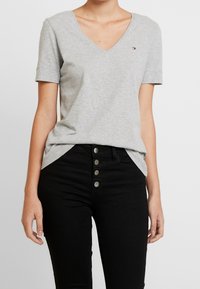 T-shirt grigio in cotone con scollatura a V e maniche corte, indossato con jeans neri a vita alta con dettagli dei bottoni. Piccolo logo sulla t-shirt.