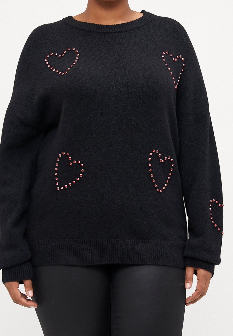 Pull noir à manches longues avec motifs de cœurs en strass rouges sur la poitrine, la taille et la manche, porté avec un pantalon noir.