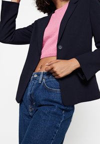 Námornický blazer s jedním knoflíkem a širokými rukávy, spárovaný s texturovaným růžovým zkráceným svetrem a džíny s vysokým pasem v modré barvě.