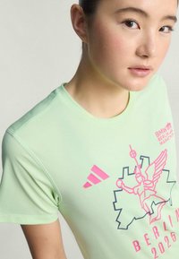 Vaaleanvihreä urheilupaita, jossa on vaaleanpunainen graafinen printti, johon kuuluu teksti "Berliini 2025" ja tyylitelty siivekäs hahmo. Paidassa on lyhyet hihat ja pyöreä pääntie.