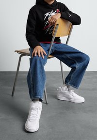 Persona che indossa sneakers bianche Nike, jeans blu e un cappuccio nero seduta all'indietro su una sedia di legno, con un pavimento grigio e una parete bianca.