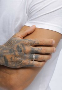 Tatuerad hand med en silverflätad ring som vilar på en böjd arm klädd i en vit kortärmad skjorta mot en neutral bakgrund.