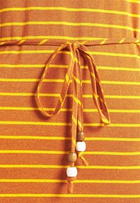 Kangaskangas, jossa on oranssia ja keltaista raidallista kuviointia, sisältää solmittavan vyötärön, jossa on koristeellisia puu- ja valkeita helmiä, korostaen tekstuuria ja värikontrasteja.