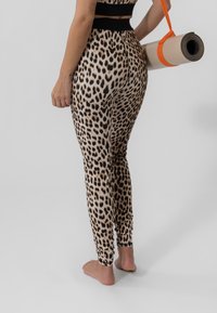 Dyreprint leggings i tan og svart, tettsittende design, jevn tekstur. Rullet yogamatte med oransje stropp holdt i venstre hånd.