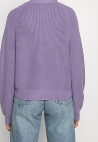 Lila gestrickter Pullover mit geripptem Saum und Raglanärmeln, kombiniert mit hellblauen Jeans. Die Textur ist dick und weich.
