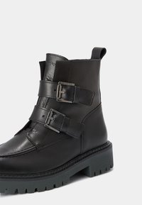 Botte cheville noire en cuir avec deux sangles ajustables à boucle, semelle épaisse à crampons et tirette à l'arrière.