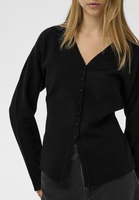 Sieviete, kas ģērbusies melnā V-veida pogājamā cardiganā ar garām piedurknēm un tumši pelēkām biksēm, stāv uz vienkārša fona.