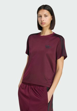 Jeune femme aux cheveux lisses et foncés portant une tenue de sport Adidas bordeaux avec des bandes noires, debout, une main dans la poche, sur un fond uni.