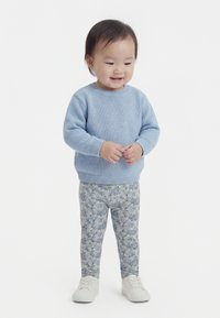 Kleinkind trägt einen hellblauen Pullover, geblümte Hose und weiße Schuhe, steht lächelnd vor einem einfarbigen weißen Hintergrund.