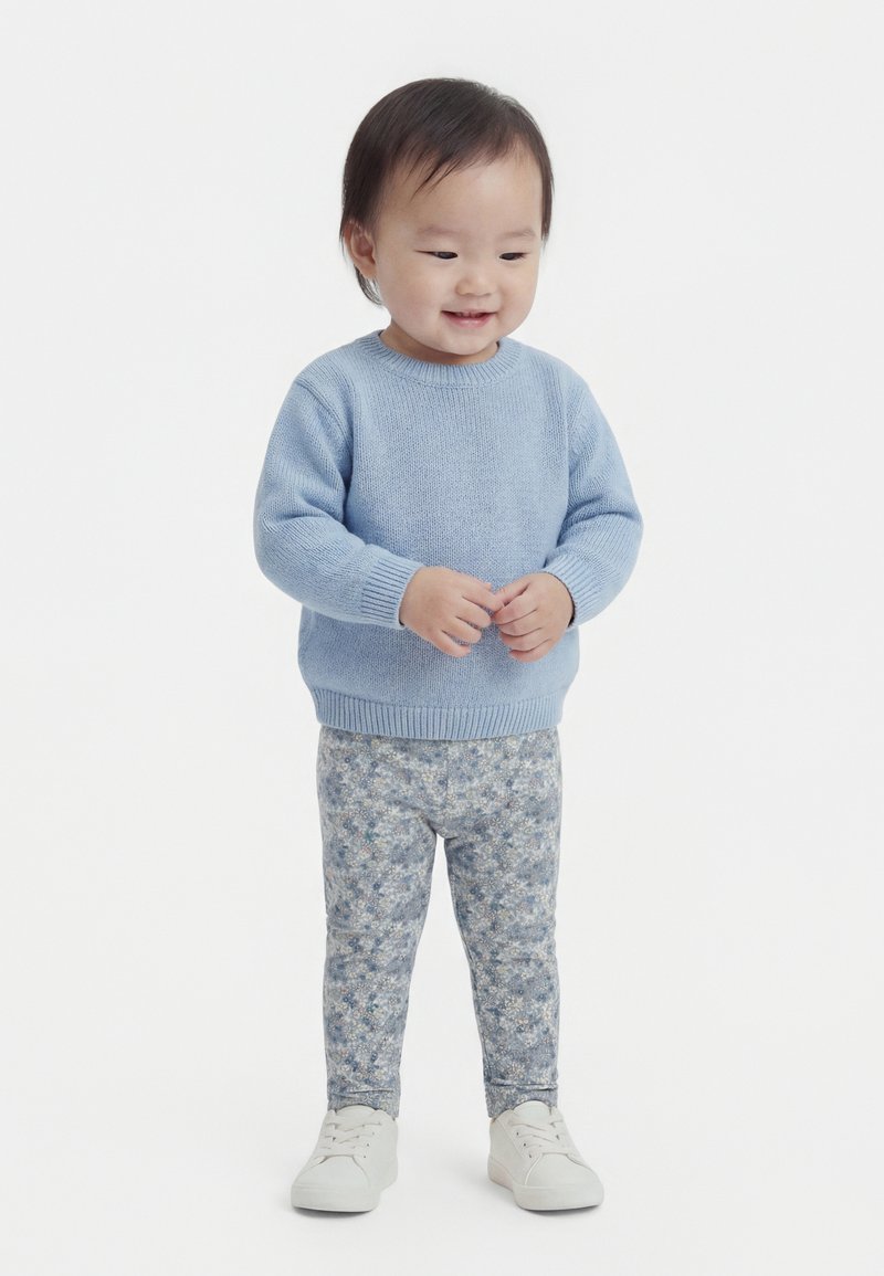 Kleinkind trägt einen hellblauen Pullover, geblümte Hose und weiße Schuhe, steht lächelnd vor einem einfarbigen weißen Hintergrund.