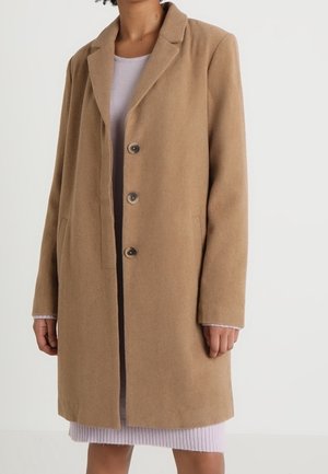 Femme portant un manteau camel mi-long à trois boutons par-dessus une robe côtelée violet clair, se tenant devant un fond blanc uni.