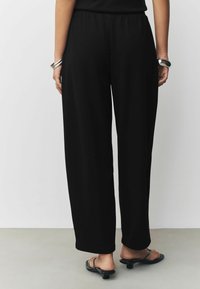 Schwarze, weit geschnittene Hosen mit elastischem Bund. Glatte Textur, knöchellang, kombiniert mit schwarzen Absatzsandalen. Einfaches und vielseitiges Design.