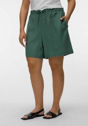 Person trägt dunkelgrüne elastische Shorts mit Kordelzug, weißes Oberteil und schwarze offene flache Sandalen, steht vor einfachem Hintergrund.