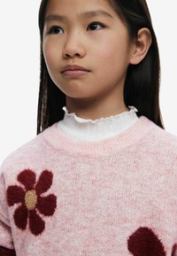 Hellrosa gestrickter Pullover mit einer dunkelroten Blumenapplikation und goldener Mitte, über einem weißen, rüschierten Rollkragenpullover getragen.