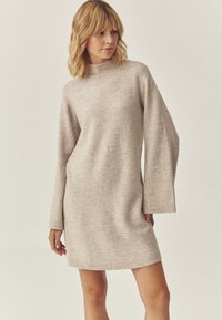 Beige kötött ruha magas gallérral, hosszú ujjakkal és bordázott mandzsettákkal. Egyszerű dizájn és sima textúra, a comb közepéig ér.