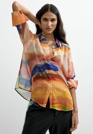 Mujer con cabello largo y oscuro lleva una blusa suelta y abotonada multicolor con patrones en naranja, azul y amarillo, posando con un brazo levantado.