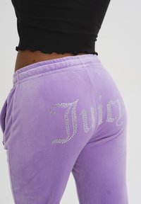 Joggings in velluto viola con logo "Juicy" in strass argentati. Presentano una vita elasticizzata e tasche laterali. Materiale morbido con finitura liscia.