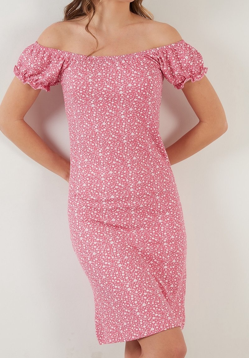 Femme portant une robe rose, épaules dégagées, longueur au genou, avec imprimé floral blanc et manches courtes bouffantes, debout les mains derrière le dos.