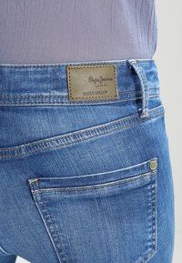 Jeans de mezclilla azul con un acabado descolorido. Presentan una etiqueta de cuero marrón con la inscripción "Pepe Jeans" y "Wiser Wash". Incluyen bolsillos traseros con detalles de costura.