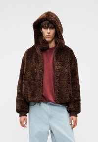 Νεαρός άνδρας με καφέ χνουδωτό μπουφάν με κουκούλα πάνω από κόκκινο πουκάμισο και ανοιχτόχρωμα μπλε τζιν, στάσιμος μπροστά σε λευκό απλό φόντο.