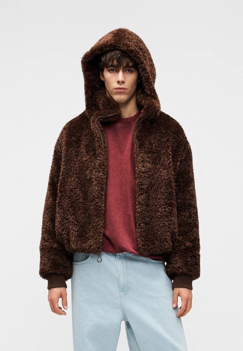 Νεαρός άνδρας με καφέ χνουδωτό μπουφάν με κουκούλα πάνω από κόκκινο πουκάμισο και ανοιχτόχρωμα μπλε τζιν, στάσιμος μπροστά σε λευκό απλό φόντο.