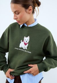 Sweatshirt vert foncé à col rond, avec une broderie blanche représentant un chien portant un bandana rouge et le texte "Toujours adorable, comme un westie."