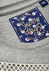 Graues T-Shirt mit einem marinefarbenen Etikett mit weißen Blumenmustern. Goldene Aufschrift "Super dry" und "OUTFITTERS." Mesh-Detail am Saum.