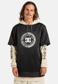Felpa con cappuccio nera e beige a maniche corte, con un grande logo DC sulla parte anteriore e dettagli stampati sulle maniche.