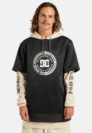 Felpa con cappuccio nera e beige a maniche corte, con un grande logo DC sulla parte anteriore e dettagli stampati sulle maniche.