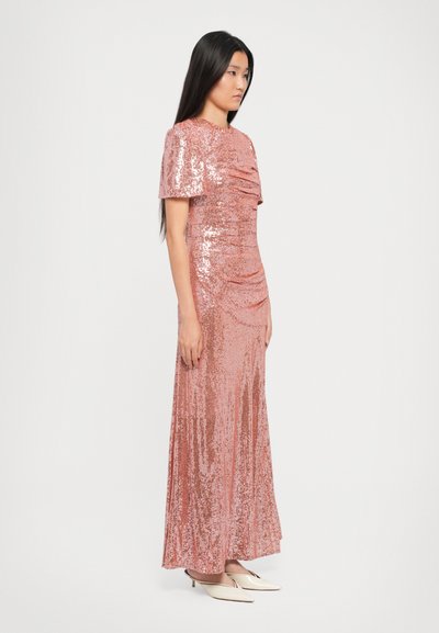 Vrouw met lang zwart haar, gekleed in een rosegouden sequinedress tot op de vloer en witte puntige hakken, zijwaarts staand op een effen achtergrond.