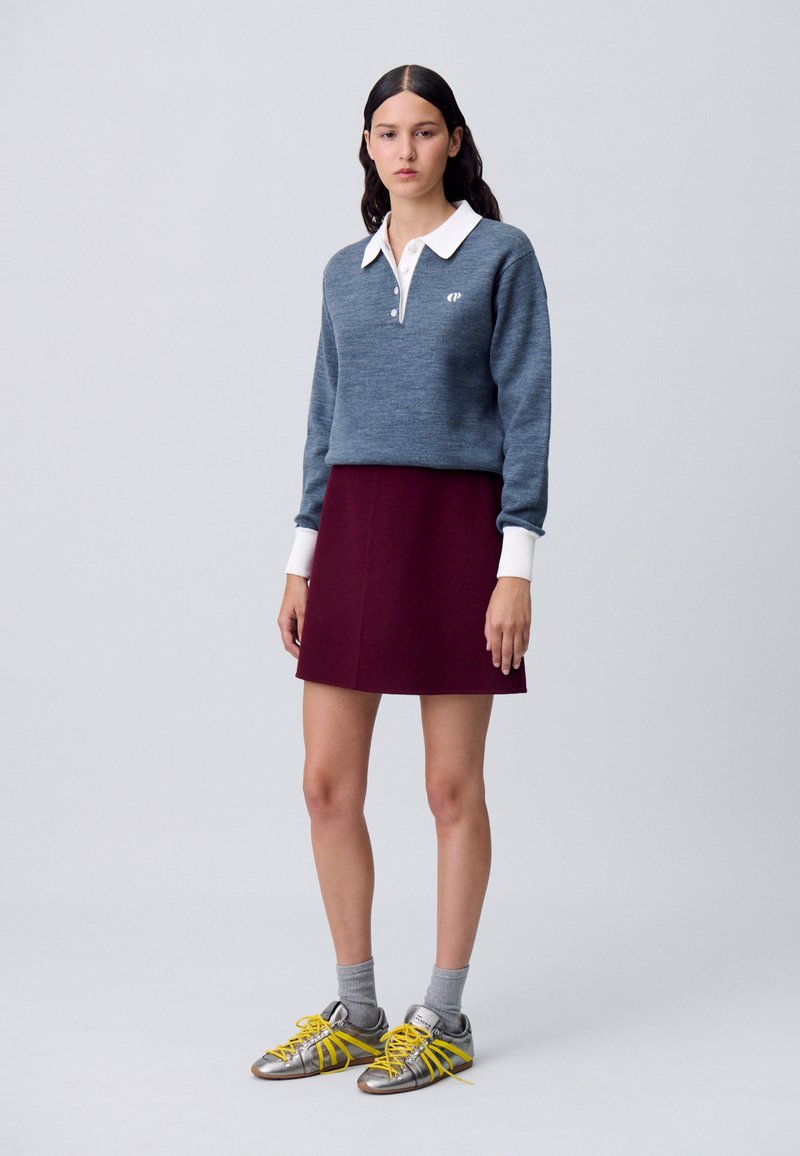 Suéter gris de punto con un cuello blanco, cuello abotonado y logo bordado, combinado con una falda burdeos. Llevado con calcetines grises y zapatillas plateadas.
